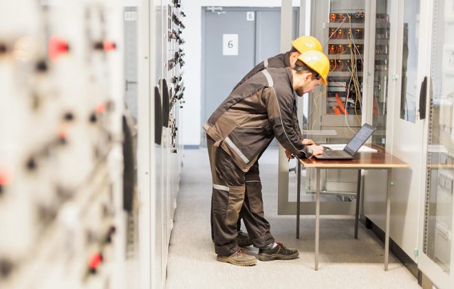Two maintenance engineers inspect relay protection system with laptop computer. Bay control unit. Medium voltage switchgear
