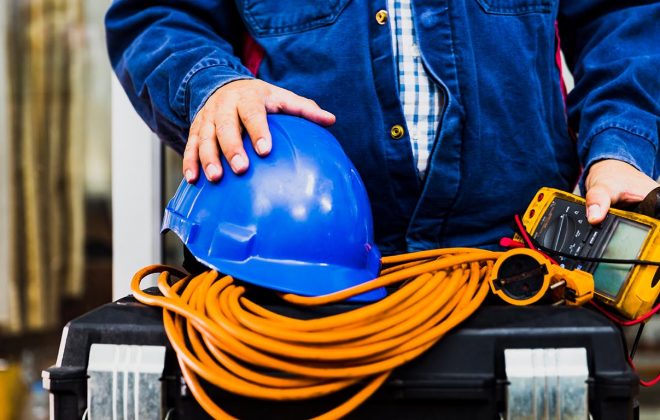 Working with safety and protection concept: Electrian engineer holding multimeter and tools in hand, standing behind the heavy duty tool box, image including power cord, Blue hard hat (helmet) and gloves.